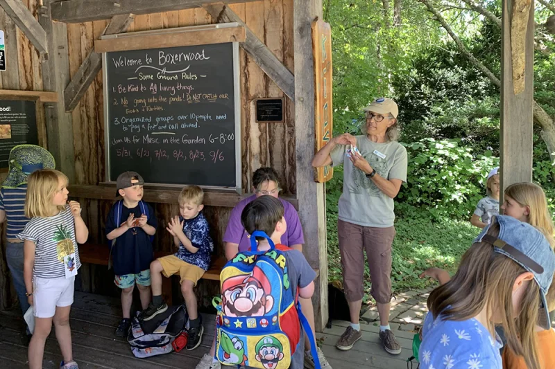 A Boxerwood educator leads a group of young children in an outdoor lesson beside a chalkboard sign