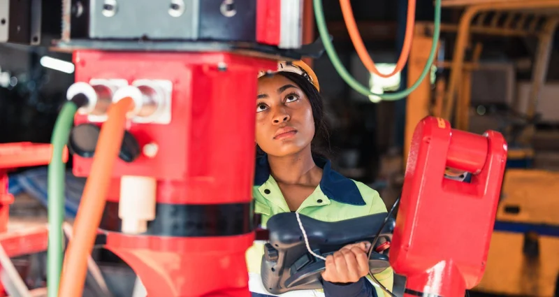 A woman in a hard hat and safety vest operates heavy industrial equipment at a skilled trades worksite