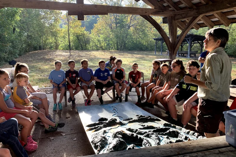 An educator addresses a group of students seated under an open-air shelter during an outdoor Boxerwood program